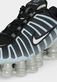 Μαύρο και γκρι αθλητικό παπούτσι Nike Shox με λευκό λογότυπο Swoosh και χαρακτηριστική παχιά, τμηματική ασημένια σόλα για απορρόφηση κραδασμών.