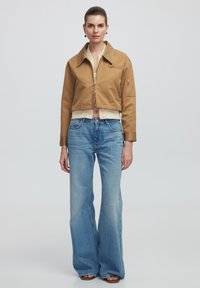 Abgeschnittene, braune Jacke mit Reißverschluss und seitlicher Tasche, kombiniert mit weit geschnittenen blauen Jeans, die leichte Verwaschungen und eine hohe Taille haben.