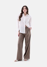 Camicia bianca con texture a pois sottili; abbinata a pantaloni ampi color marrone chiaro. Modello che indossa sandali intrecciati di colore chiaro.