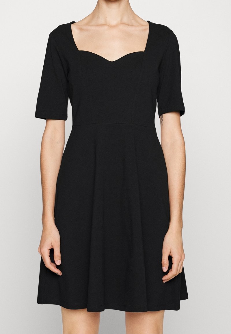 Femme portant une robe noire à manches courtes avec un corsage ajusté et un décolleté en cœur, debout devant un fond clair uni.