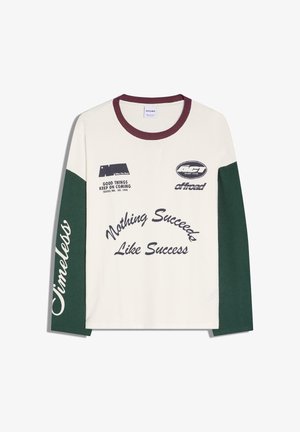 Wit shirt met lange mouwen, groene mouwen, een bordeauxrode kraag en tekst waaronder "Nothing Succeeds Like Success" en "Timeless" op de mouw.