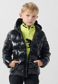 Chaqueta acolchada negra con acabado brillante, cierre de cremallera y capucha. Lleva puesta sobre un suéter con cremallera de color verde brillante que presenta un patrón negro.