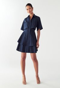 Robe bleu marine avec design à col, fermeture éclair à l'avant, manches courtes et jupe à volants. Le tissu lisse et l'ourlet superposé créent une silhouette structurée.