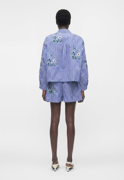 Personne debout de dos portant une chemise oversize rayée bleu et blanc et un short assorti avec des fleurs blanches brodées, sandales blanches à talons.
