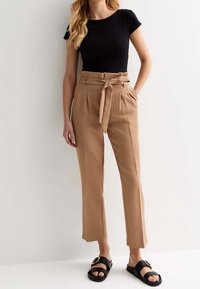 Zwarte strakke t-shirt, beige high-waisted broek met een strikriem, cropped lengte, gladde stof en zwarte sandalen met een metalen accent.