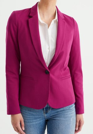 Kvinde iført en tætsiddende magenta blazer med en knap over en hvid skjorte og blå jeans mod en ensfarvet baggrund.