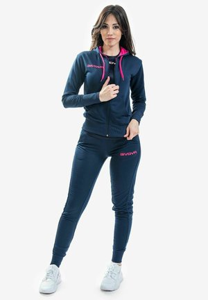 Ung kvinde i navyblå og pink Givova træningsdragt og hvide sneakers, der står mod en ensfarvet hvid baggrund.