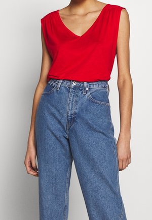 Haut rouge sans manches avec un col en V, associé à un jean en denim bleu clair taille haute. Le haut présente un léger froncé aux épaules et une texture lisse.