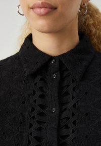 Črna majica z teksturirano tkanino in izrezljanimi vzorci, s pointed ovratnikom in gumbi spredaj.