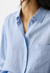 Ljusblå linnekorta med krage, har en enskild bröstficka och vita knappar, som visar ett slätt, strukturerat tyg.