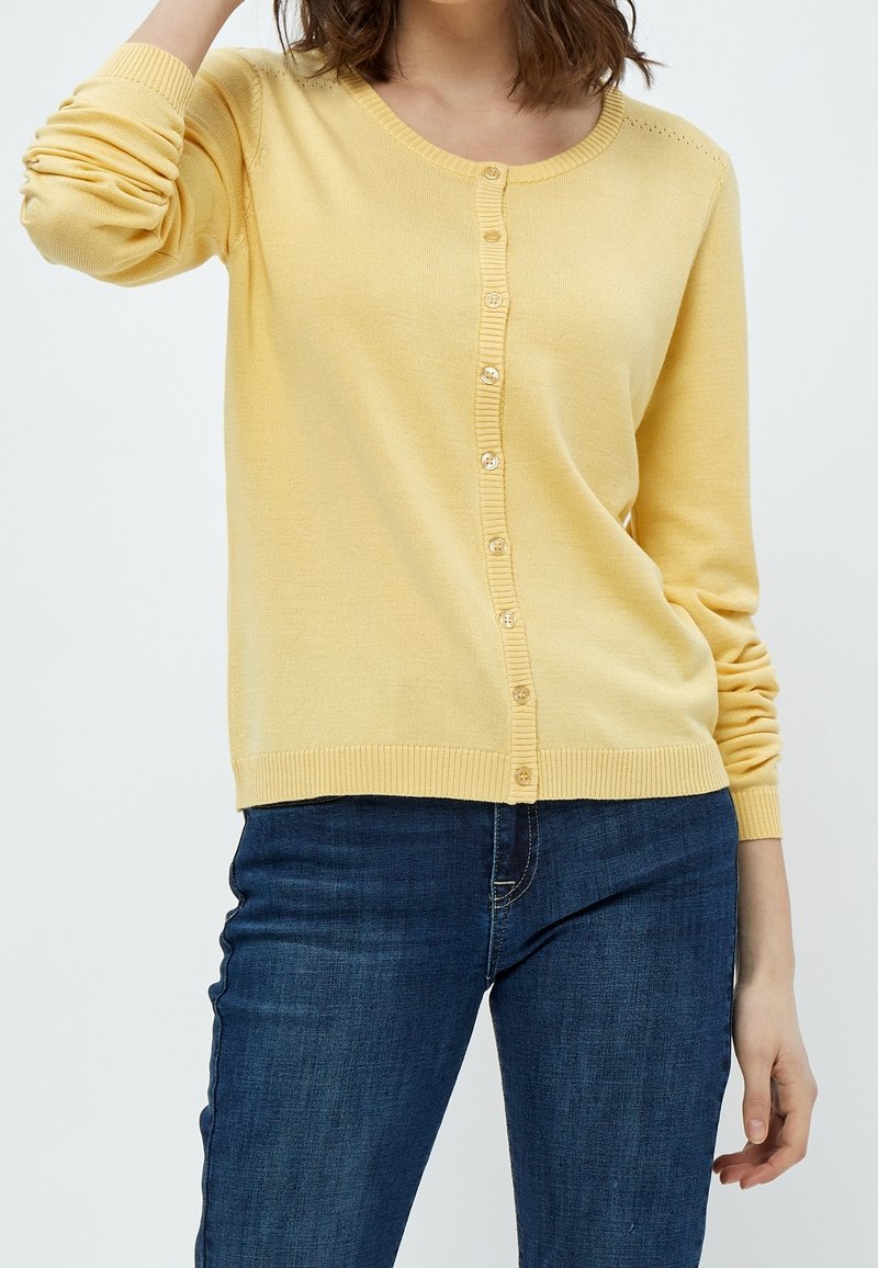 Gulstickad cardigan med rund hals och ribbad fåll. Har knappstängning och texturerad detalj på axlarna. Bärs tillsammans med blå jeans.