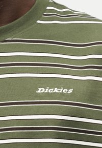 Κοντινό πλάνο ενός πράσινου και λευκού ριγέ πουκάμισου με το λογότυπο "Dickies" κεντημένο στο στήθος με άσπρα γράμματα.