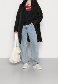 Musta eristämätön pitkä takki hupulla, päällä tummansininen ja punainen kuvioitu neulepaita. Vaaleansiniset leveälahkeiset farkut ja valkoinen laukku täydentävät asun.