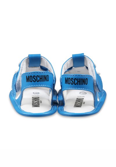 Sandales pour bébé bleues avec une finition brillante, des sangles en velcro ajustables et une marque "Moschino" sur le dessus. Doublure intérieure blanche lisse.