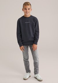 Usmievajúci sa chlapec stojí, oblečený v čiernej mikine s nápisom "Blue Ridge Denim", šedých džínsoch a bielych teniskách na jednoduchom pozadí.