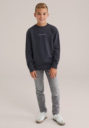 Ragazzo sorridente in piedi, che indossa una felpa nera con la scritta "Blue Ridge Denim", jeans grigi e sneakers bianche su uno sfondo semplice.