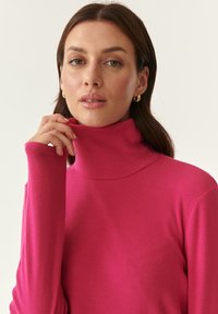Maglione a collo alto a coste, aderente e di un vivace colore rosa. Tessuto morbido, maniche lunghe e collo alto. Orecchini dorati visibili nel ritratto.