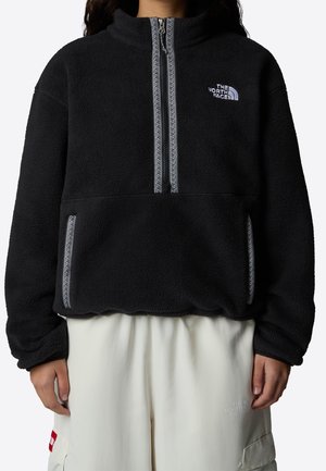 Osoba ubrana w czarny polar z półzamkiem i białym logo oraz jasne spodnie z bocznymi kieszeniami i subtelnym brandingiem.