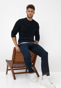 Maglione nero lavorato a maglia, jeans scuri e sneaker bianche. Modello seduto su una sedia di legno con design della seduta intrecciato. Aspetto semplice e moderno.