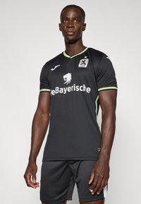 Juoda sportinė apranga su trumpomis rankovėmis, dekoruota įvairiaspalviu apvadu, liūto logotipu ir rėmėjo tekstu. Derinama su juodais šortais, tekstūruota medžiaga.