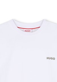 Camiseta blanca de algodón con cuello redondo, mangas cortas y un logo gris de "HUGO" en el lado izquierdo del pecho. La etiqueta interior es roja.