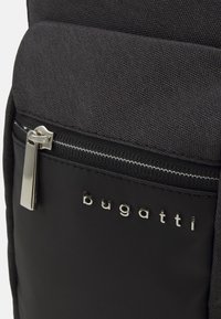 Zaino in tessuto nero caratterizzato da una tasca frontale liscia e opaca con una cerniera argentata e il logo in rilievo "bugatti" in lettere metalliche.