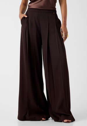 Personne portant un pantalon plissé large marron foncé avec des poches, debout avec une main dans la poche et les pieds visibles en sandales.