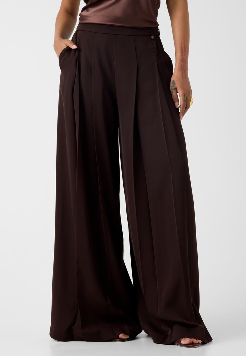 Personne portant un pantalon plissé large marron foncé avec des poches, debout avec une main dans la poche et les pieds visibles en sandales.