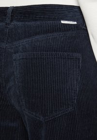 Námořnické kordové kalhoty s vertikálními pruhy, s jednou zadní kapsou a malou nášivkou s nápisem "Marc O'Polo BENIN." Měkká textura a upravený střih.