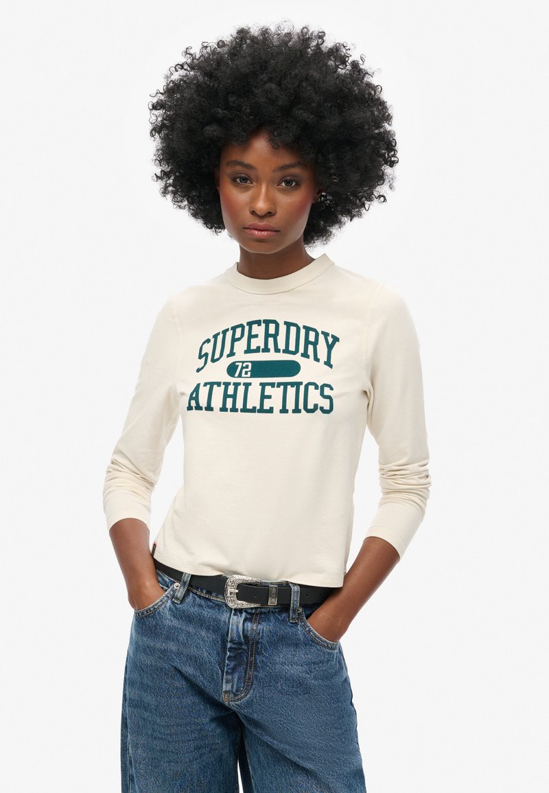 Kremowa koszulka z długim rękawem z napisem "SUPERDRY 72 ATHLETICS" w ciemnozielonym kolorze. Połączona z granatowymi dżinsami z wysokim stanem i czarnym paskiem.