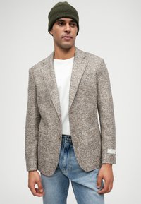Men's beige textured blazer with a notched collar, single button closure, patch pockets, and label detail on the sleeve, worn over a white shirt.
