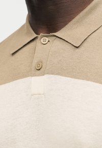 Gros plan sur une personne portant un polo en maille beige et blanche avec deux boutons beiges fermés au niveau du col.