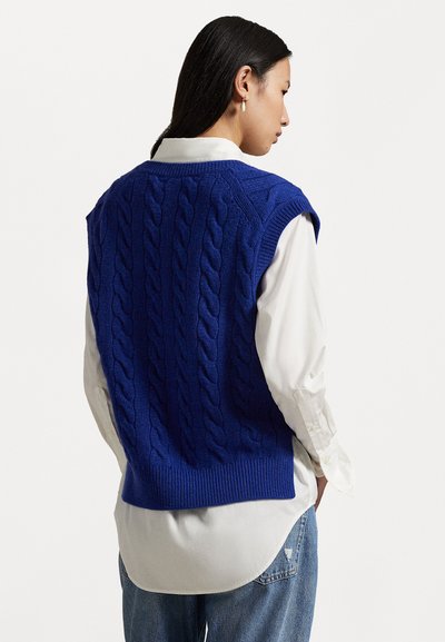 Blue knitted sweater vest with cable pattern, ribbed hem, and sleeveless design, layered over a white button-up shirt. Casual fit.