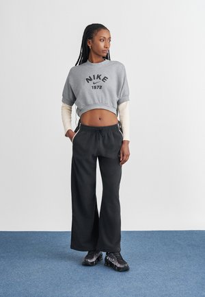 Sweatshirt cinzento cropped com o texto "NIKE 1972", combinado com calças de fato largas pretas. A modelo usa sapatilhas desportivas pretas com detalhes prateados.
