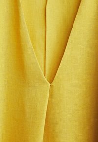 Tissu jaune avec un tissage texturé, présentant un décolleté en V et un détail de couture centrale. Léger et tombe en douceur.