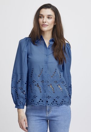 Blaue Bluse mit Puffärmeln, die am Saum und an den Ärmeln mit ausgeschnittenen Blumendrucken versehen ist, aus leichtem Baumwollstoff gefertigt.