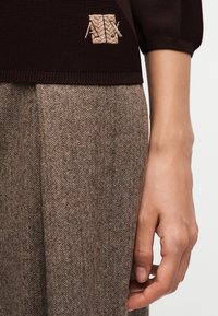 Gros plan d'une main à côté d'un pantalon marron à motif chevrons et d'un haut en maille foncé avec un logo "A|X" beige brodé.