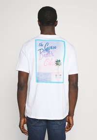 Weißes Baumwoll-T-Shirt mit einem bunten Strandclub-Motiv auf der Rückseite, das Palmen, den Ozean und stilisierte Schriftzüge in Pink und Blau zeigt.