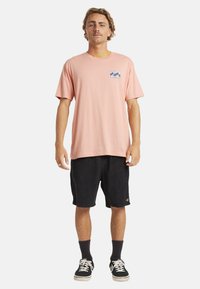 Różowa bawełniana koszulka z krótkim rękawem i falistym wzorem na lewej piersi, zestawiona z czarnymi szortami z sztruksu i czarnymi trampkami.