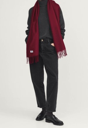 Personne portant un jean noir, un pull à col roulé gris foncé, une longue écharpe bordeaux avec des franges, et des mocassins en cuir noirs, debout les mains dans les poches.