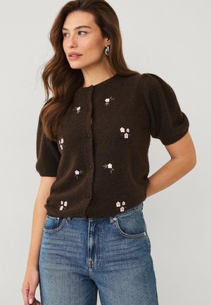 Frau trägt dunkelbraunen kurzärmeligen Cardigan mit kleiner rosa Blumenstickerei und blaue Jeans, posiert mit der Hand hinter dem Rücken.