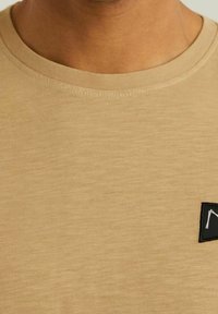 Béžové bavlněné tričko s kulatým výstřihem, které má na přední straně malou černou nášivku s bílým logem. Měkká textura, minimalistický design.