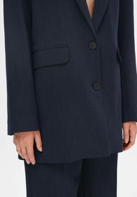 Marineblazer met twee zwarte knopen, één borstzak en een gladde textuur. Heeft een gestructureerde pasvorm en zichtbare stikdetails.