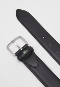 Zwarte leren riem met een zilveren rechthoekige gesp, voorzien van een Lacoste-logo en stikseldetails op de riem. Enkele pin-sluiting.