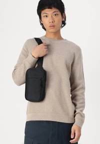 Geantă neagră de umăr cu buzunare cu fermoar, textură netedă și formă rectangulară, purtată peste un pulover tricotat bej.