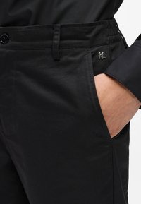 Melnas bikses, kas izgatavotas no gludas auduma, ar jostu, kurai ir elastīga daļa, sānu kabatām un nelielu metāla logotipa detaļu.