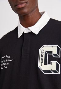 Schwarzes Polohemd mit weißem Kragen, bestickt mit dem "CRHT"- und "C"-Logo in Schwarz und Beige. Auf der linken Seite steht der Text "Million To Millions."