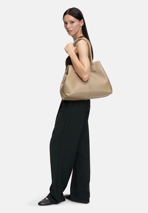 Borsa tote in pelle beige con due manici corti, struttura morbida e cuciture minime, tenuta da un modello vestito con pantaloni neri.