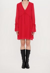 Rode, vloeiende jurk met lange, gerimpelde mouwen, een V-hals en een strikdetail bij de hals. Zwarte kniehoge laarzen maken de outfit compleet.