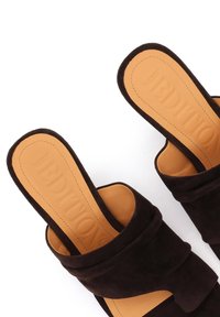 Schwarze Slip-On-Sandalen mit breiten Wildleder-Überkreuzriemen und braunen, gepolsterten Innensohlen, geprägt mit dem Markennamen, auf weißem Hintergrund.